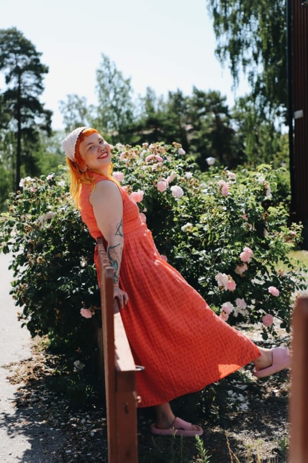 Punahiuksinen tyttö punertavassa pitkässä mekossa seisoo puutarhassa